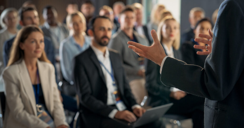 Back View Of Environment Specialist Gesturing And Presenting To Audience Of Diverse Business Leaders. Male And Female Attendees Listening To Inspirational Speech On International Economic Forum.